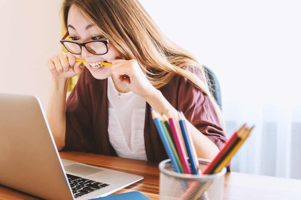 A woman sits in front of her laptop wearing glasses and biting a pencil
